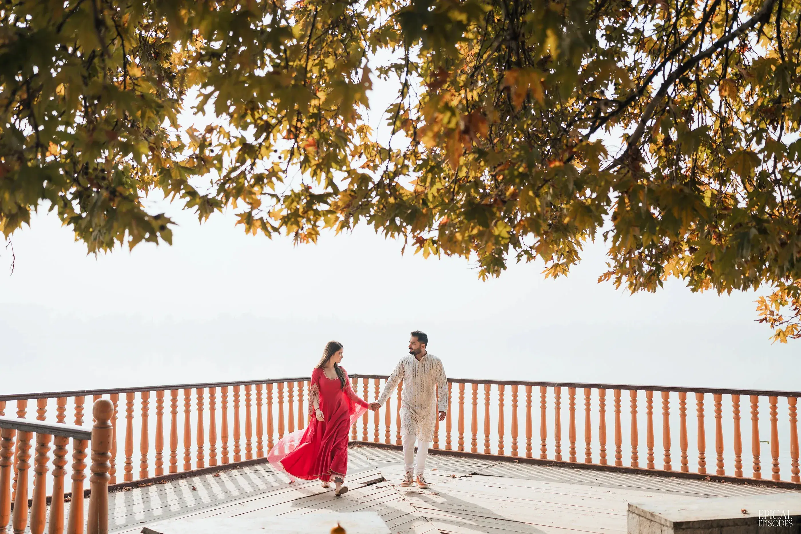 A couple walking hand in hand along a balcony beneath autumn leaves